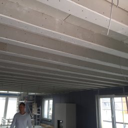 Damian Paszenda - Montaż płyt karton-gips na suficie w trakcie prac wykończeniowych. Widoczne profile i mocowania. W tle okna i fragmenty rusztowania.