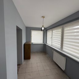 PasZenda - uslugi budowlane - Wnętrze pokoju po malowaniu ścian na szaro, z widoczną szafką, grzejnikiem i oknami z żaluzjami. Na suficie wisi lampa. Widoczne ślady po remoncie na podłodze.