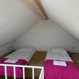 PasZenda - uslugi budowlane - Poddasze z dwoma łóżkami na drewnianej platformie, białymi ścianami i skośnym sufitem. Łóżka mają białą pościel, fuksjowe narzuty i złożone ręczniki. Widoczna drewniana belka i biała balustrada.