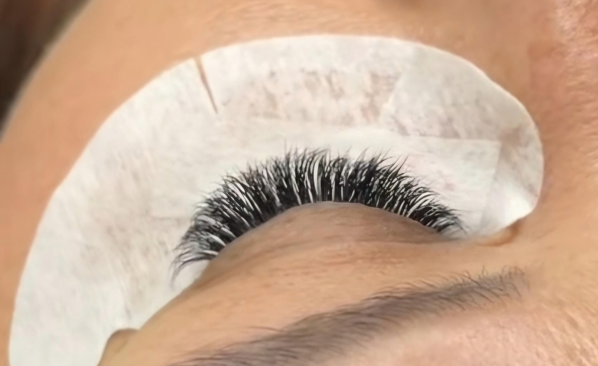 Zbliżenie na oko z gęstymi, czarnymi, przedłużonymi rzęsami. Biały płatek kosmetyczny pod dolną powieką. Widoczne naturalne brwi.