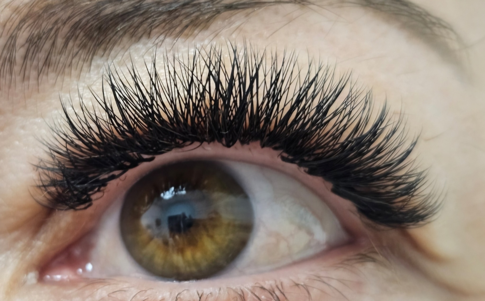 Zbliżenie na oko z przedłużonymi, gęstymi, czarnymi rzęsami. Widoczny brązowy kolor tęczówki i delikatny makijaż oka. Efekt profesjonalnej stylizacji rzęs.