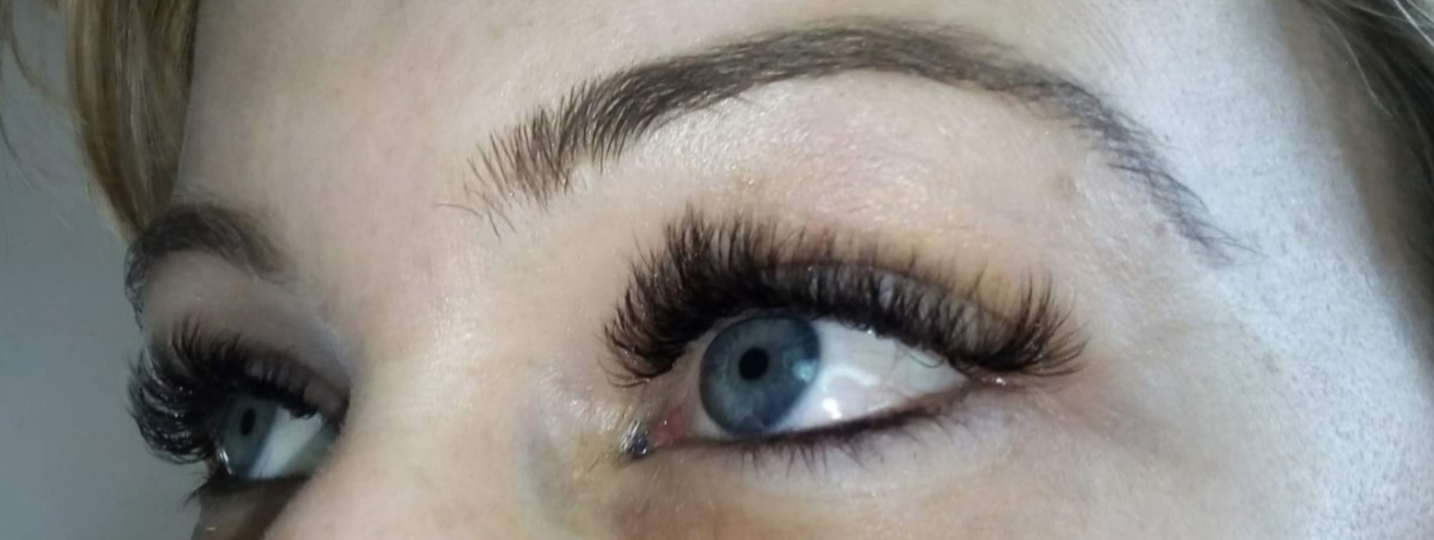 Zbliżenie na oko z gęstymi, profesjonalnie przedłużonymi rzęsami. Widoczny delikatny makijaż brwi i błękitna tęczówka. Naturalne światło.