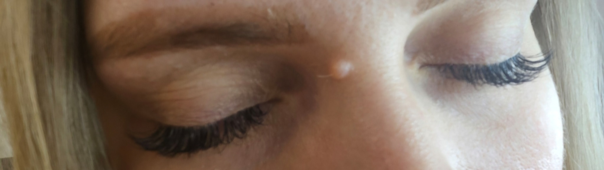 Zbliżenie na oko z przedłużonymi rzęsami, widoczny fragment blond włosów. Delikatny makijaż podkreśla spojrzenie. Na twarzy widoczny mały pieprzyk.