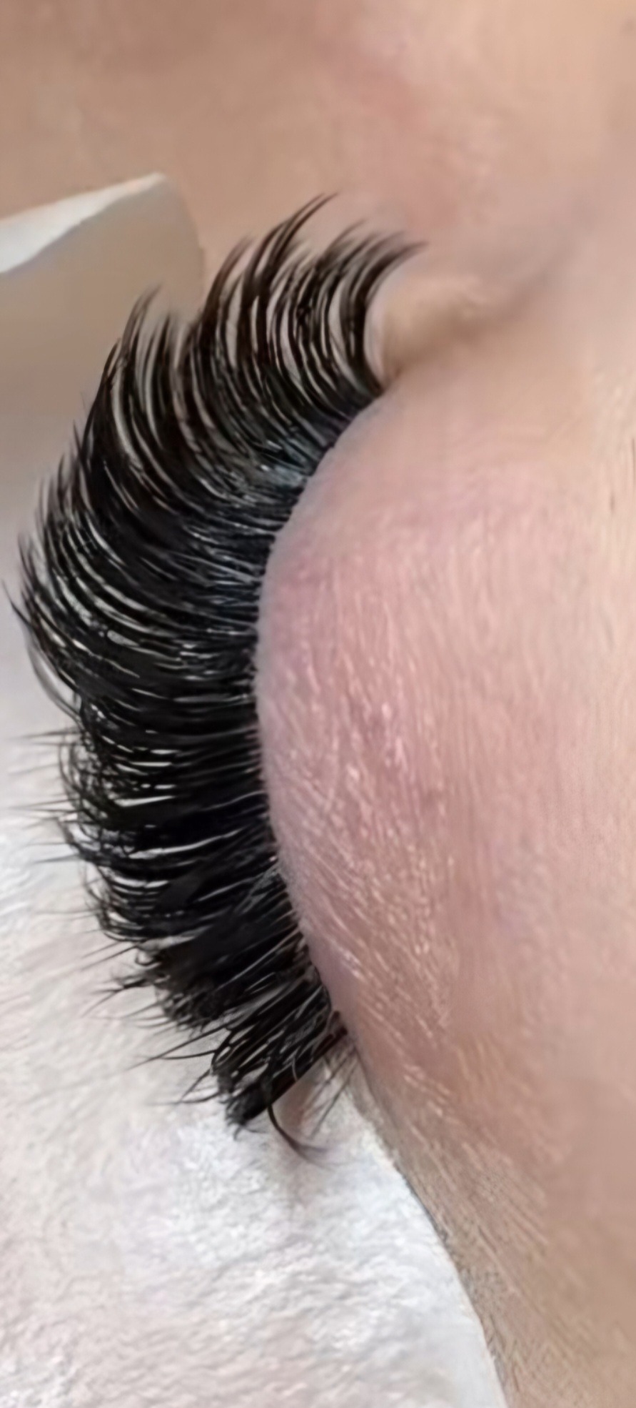 Zbliżenie na oko z gęstymi, czarnymi, profesjonalnie przedłużonymi rzęsami. Rzęsy wyraźnie oddzielone, tworzące efekt wachlarza. Widoczna skóra powiek i fragment białego podłoża.