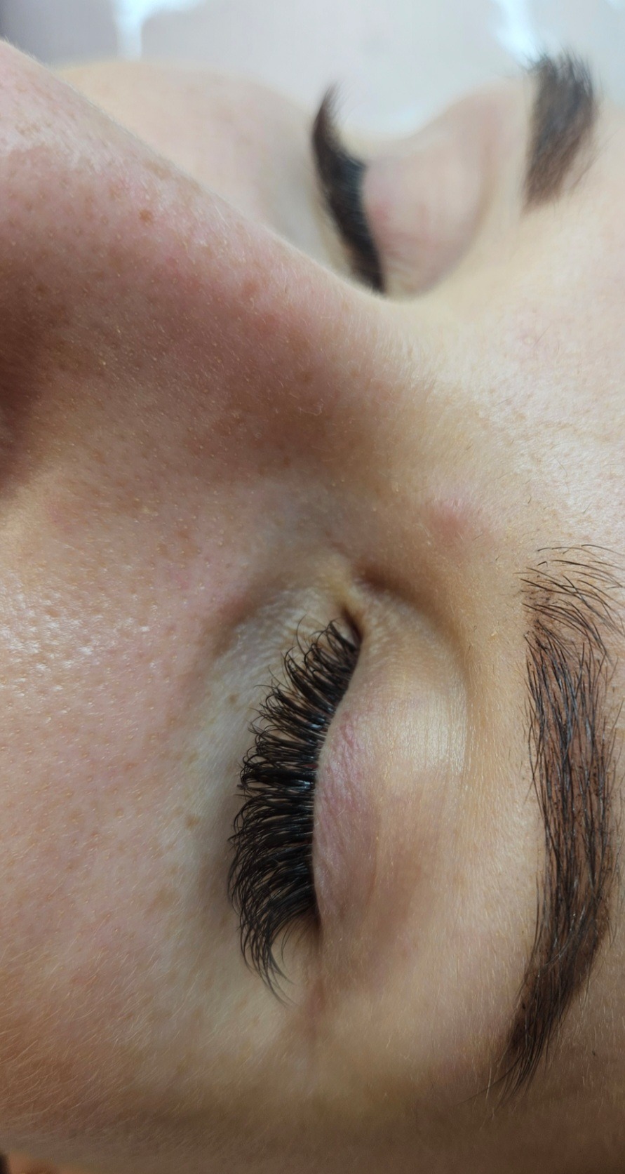 Zbliżenie na oko z przedłużonymi, czarnymi rzęsami i brwiami po zabiegu kosmetycznym, widoczne piegi na skórze.
