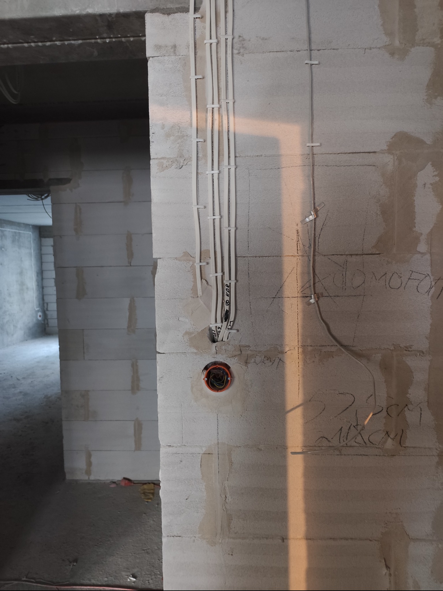 Instalacja elektryczna w surowym wnętrzu: kable prowadzone po ścianie z betonu komórkowego, widoczne puszki instalacyjne i napisy na ścianie. Etap prac wykończeniowych.