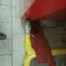 Bartosz Jaworski Usługi Ogólnobudowlane - Żółta rura gazowa z zaworem pod czerwonym piecem, widok z dołu, szare płytki w tle, fragment buta w lewym dolnym rogu kadru.