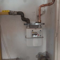 Bartosz Jaworski Usługi Ogólnobudowlane - Zamontowany gazomierz z żółtą rurą doprowadzającą gaz i miedzianą rurą odprowadzającą, szara ściana w tle. Widoczne złącza i elementy mocujące.