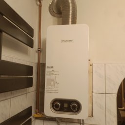 Bartosz Jaworski Usługi Ogólnobudowlane - Biały piec gazowy Junkers na ścianie z beżowymi płytkami, obok grzejnik drabinkowy i rura miedziana. Widoczne elementy instalacji i odbicie osoby w lustrze.