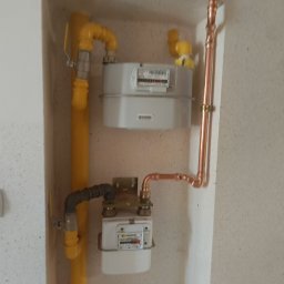 Bartosz Jaworski Usługi Ogólnobudowlane - Dwa gazomierze wnęce: górny - nowy, szary, dolny - starszy. Żółte rury gazowe i miedziane rury. Biały dzwonek po lewej. Instalacja gazowa.