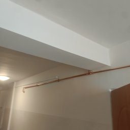 Bartosz Jaworski Usługi Ogólnobudowlane - Miedziana rura instalacji biegnąca wzdłuż ściany korytarza, zamocowana do sufitu. Widoczne łączenia rur i elementy mocujące.