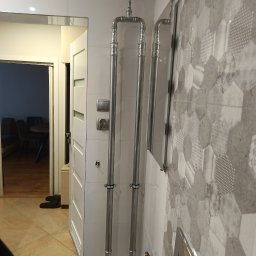 Bartosz Jaworski Usługi Ogólnobudowlane - Chromowane rury instalacji grzewczej w łazience z heksagonalnymi płytkami, widok na otwarte drzwi i fragment pomieszczenia z ciemnymi krzesłami.