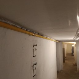 Bartosz Jaworski Usługi Ogólnobudowlane - Żółta rura gazowa biegnąca wzdłuż sufitu piwnicy, widoczne ślady po kuciu w ścianie, w tle osoba w przejściu. Instalacja gazowa po remoncie.