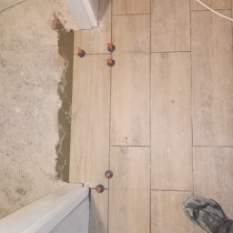 Bartosz Jaworski Usługi Ogólnobudowlane - Układanie podłogi z płytek imitujących drewno, z użyciem poziomujących klipsów. Widoczny fragment stopy w roboczym obuwiu i kabel zasilający.