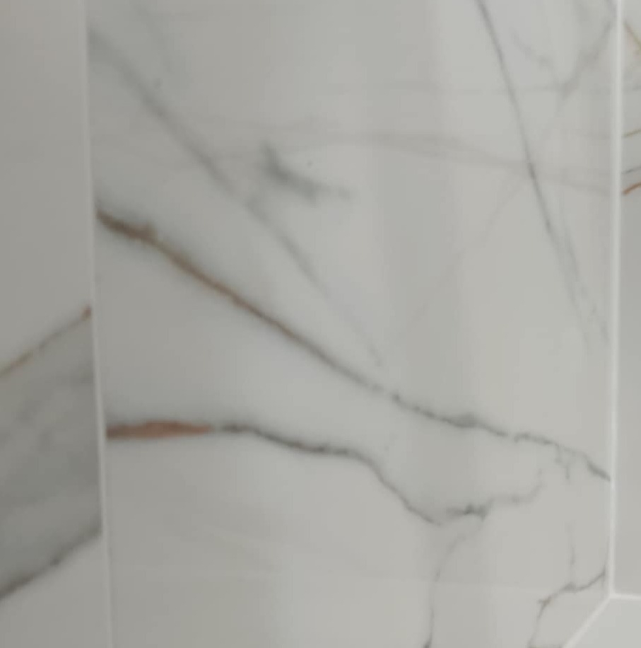 Białe płytki imitujące marmur z subtelnymi, szarymi i złotymi żyłkami, tworzące eleganckie wykończenie ściany w łazience. Widoczne fugi.