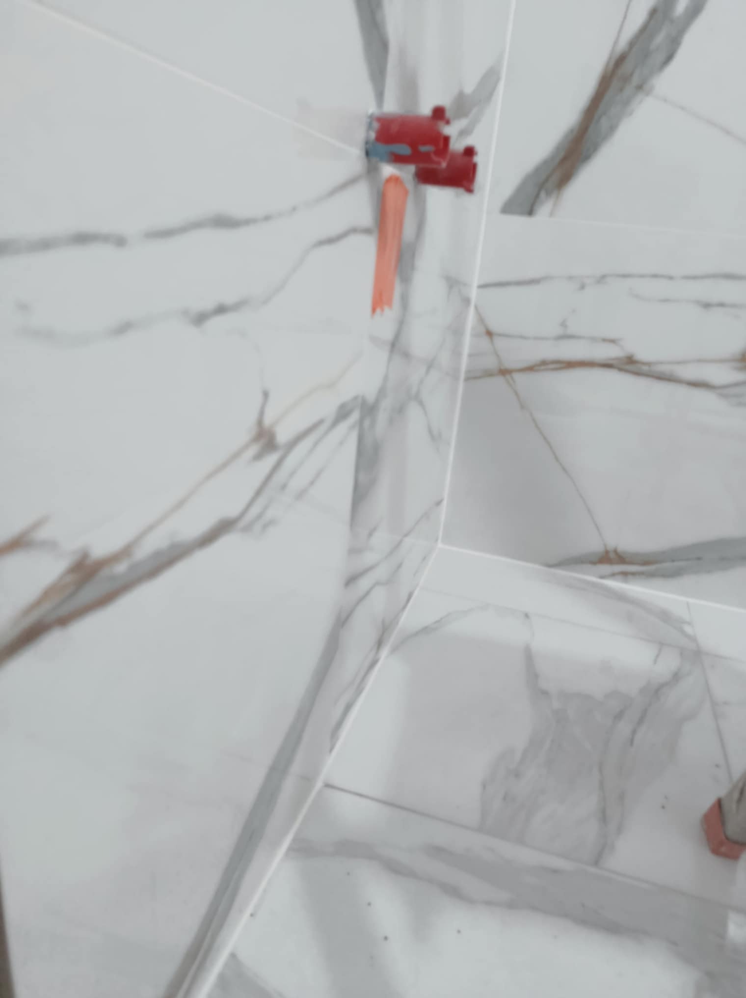 Ściana i podłoga wyłożone białymi płytkami imitującymi marmur, z widoczną czerwoną armaturą i fragmentem pomarańczowej rurki, prawdopodobnie w trakcie montażu.