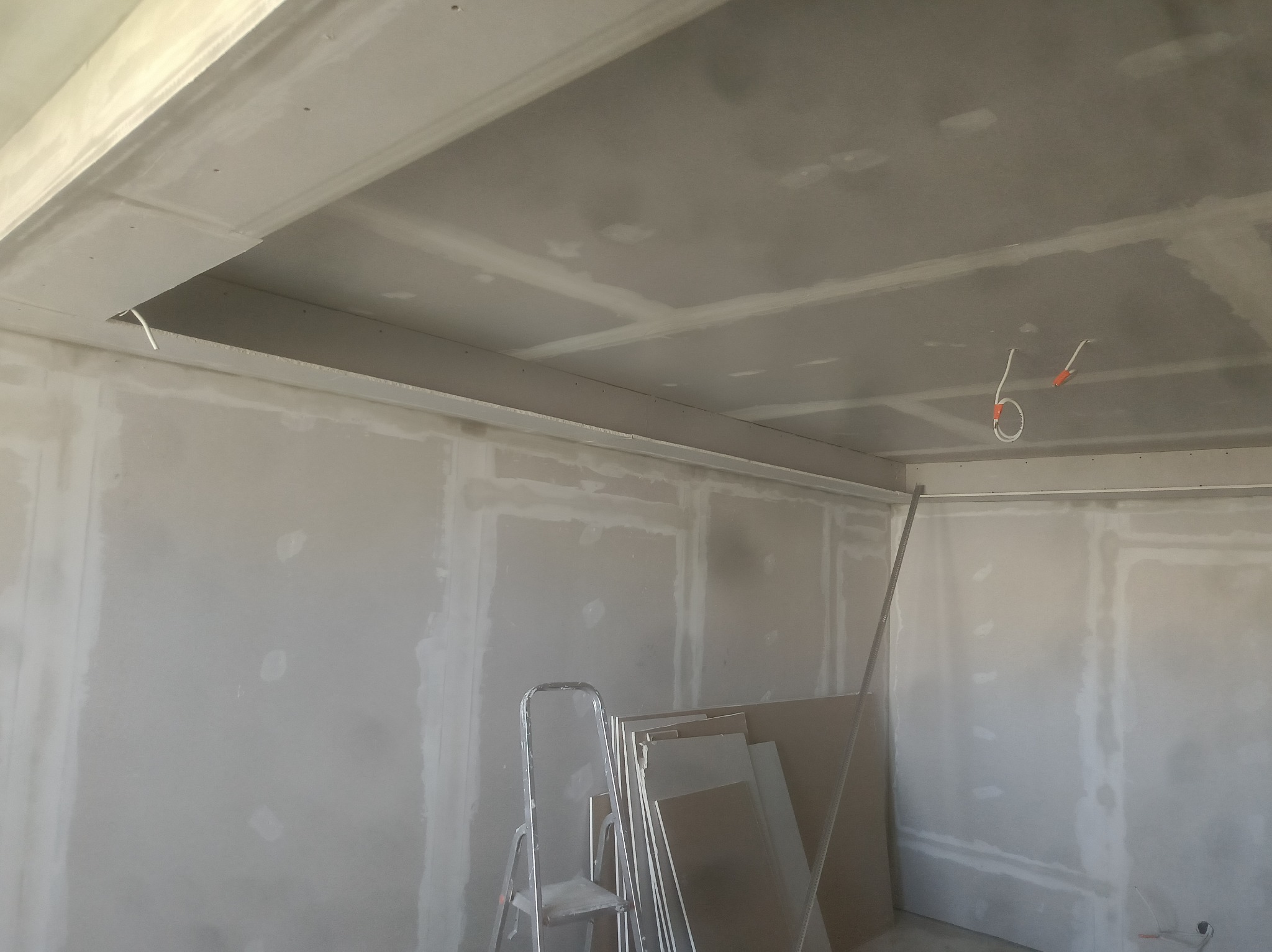 Ściany i sufit w trakcie wykańczania płytami gipsowo-kartonowymi, widoczne kable elektryczne. Aluminiowa drabina i płyty gipsowe przy ścianie.