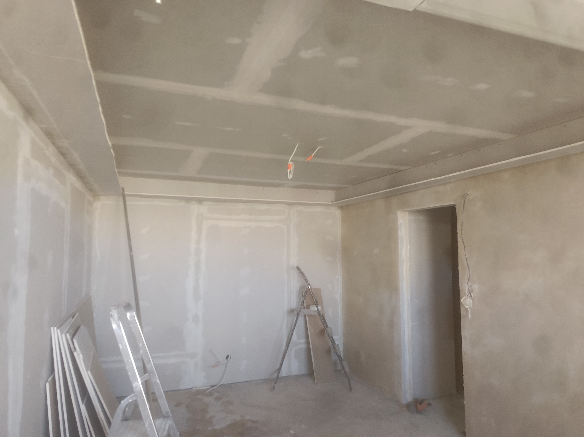 Wnętrze w trakcie remontu: ściany i sufit pokryte płytami gipsowo-kartonowymi, widoczne drabiny i materiały budowlane, przygotowanie do dalszych prac wykończeniowych.