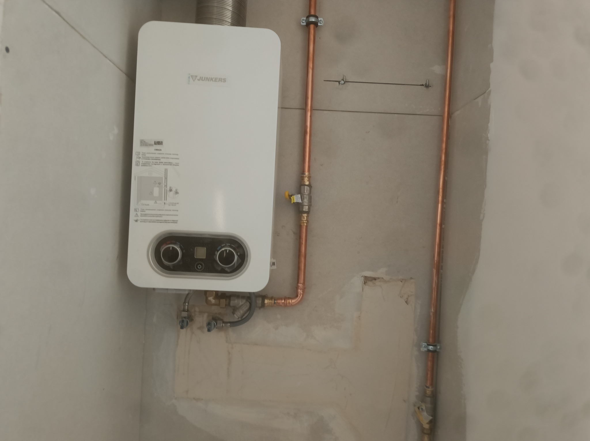 Biały piec gazowy Junkers zamontowany na ścianie z miedzianymi rurami w Chorzowie. Widoczne przyłącza i fragment komina. Ściana w surowym stanie.