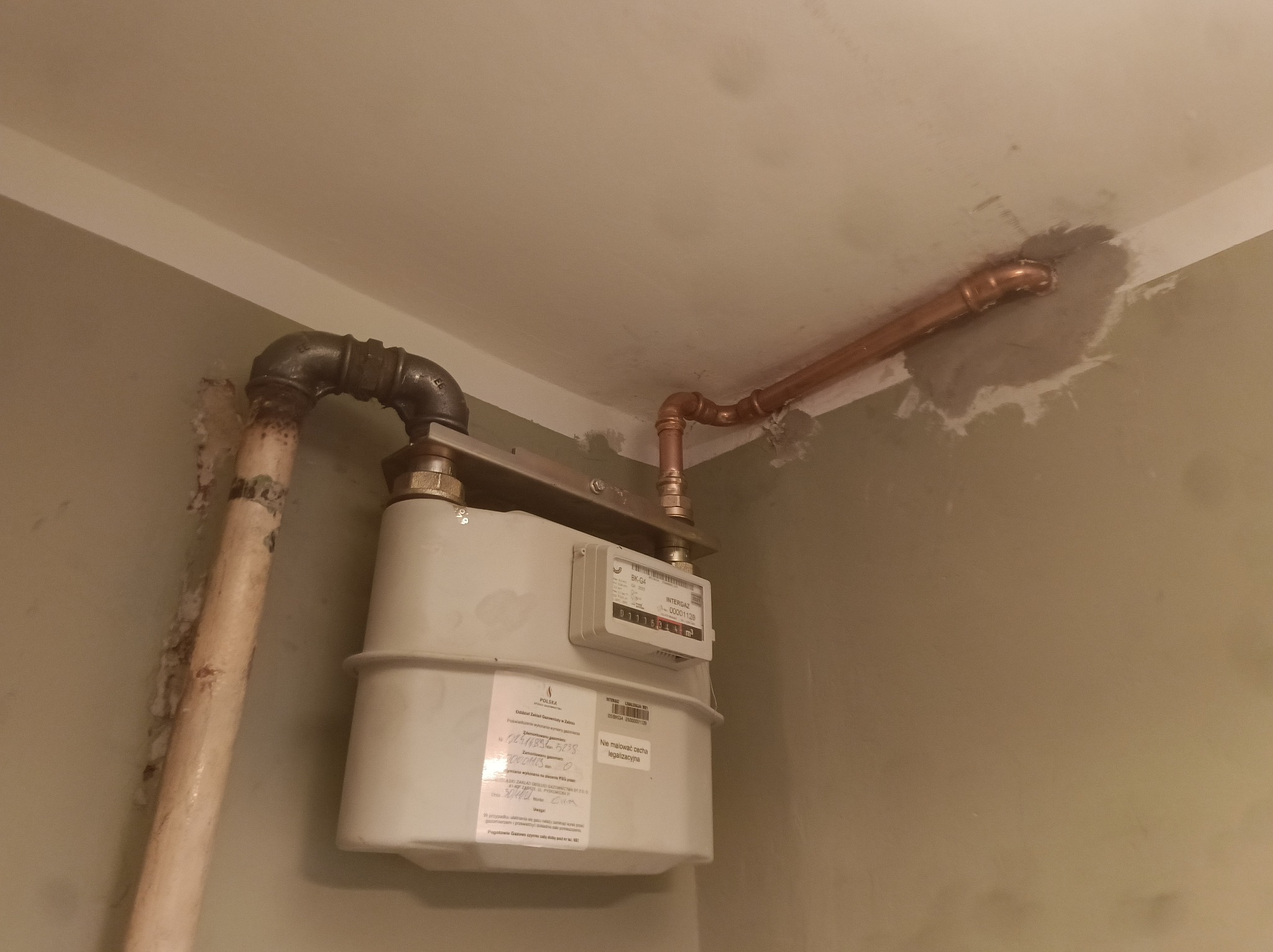 Zamontowany gazomierz z rurami miedzianymi i stalowymi na ścianie w pomieszczeniu. Widoczne ślady po poprzedniej instalacji. Ściany w odcieniach beżu i zieleni.
