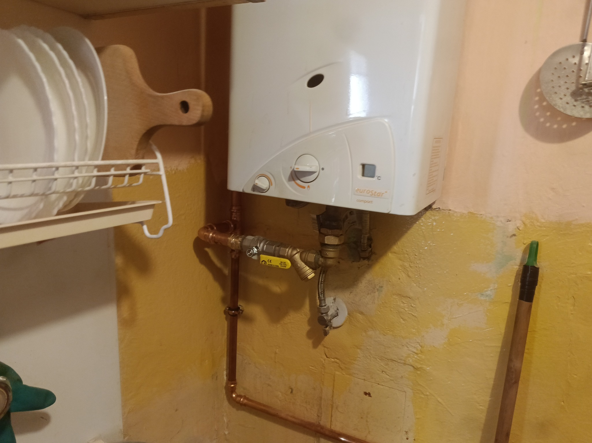 Biały piec gazowy Eurostar Compact zamontowany na żółtej ścianie, z widocznymi miedzianymi rurami i zaworami. Obok suszarka do naczyń z talerzami i drewniana deska.