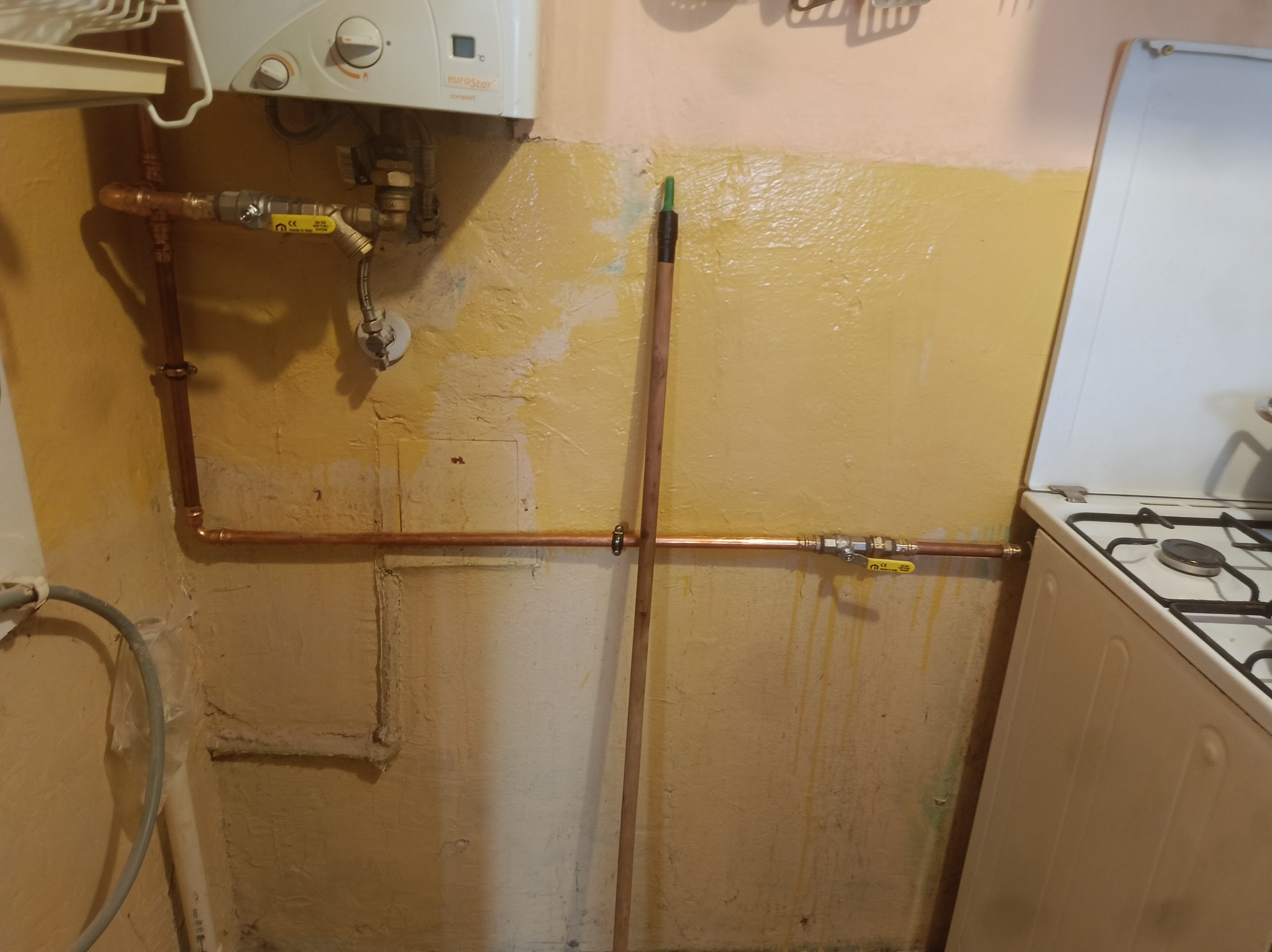 Miedziane rury gazowe z zaworami, zamontowane na nierównej, żółtej ścianie w kuchni, obok pieca gazowego i pod bojlerem. Widoczne złącza i mocowania.