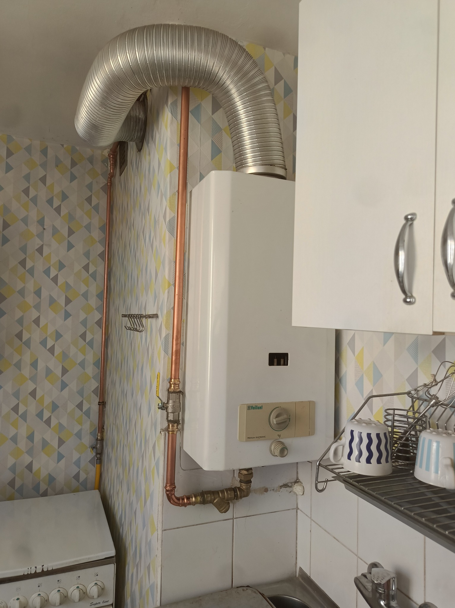 Biały piec gazowy Vaillant z miedzianymi rurami i giętką rurą odprowadzającą spaliny w kuchni z geometryczną tapetą i szafkami.