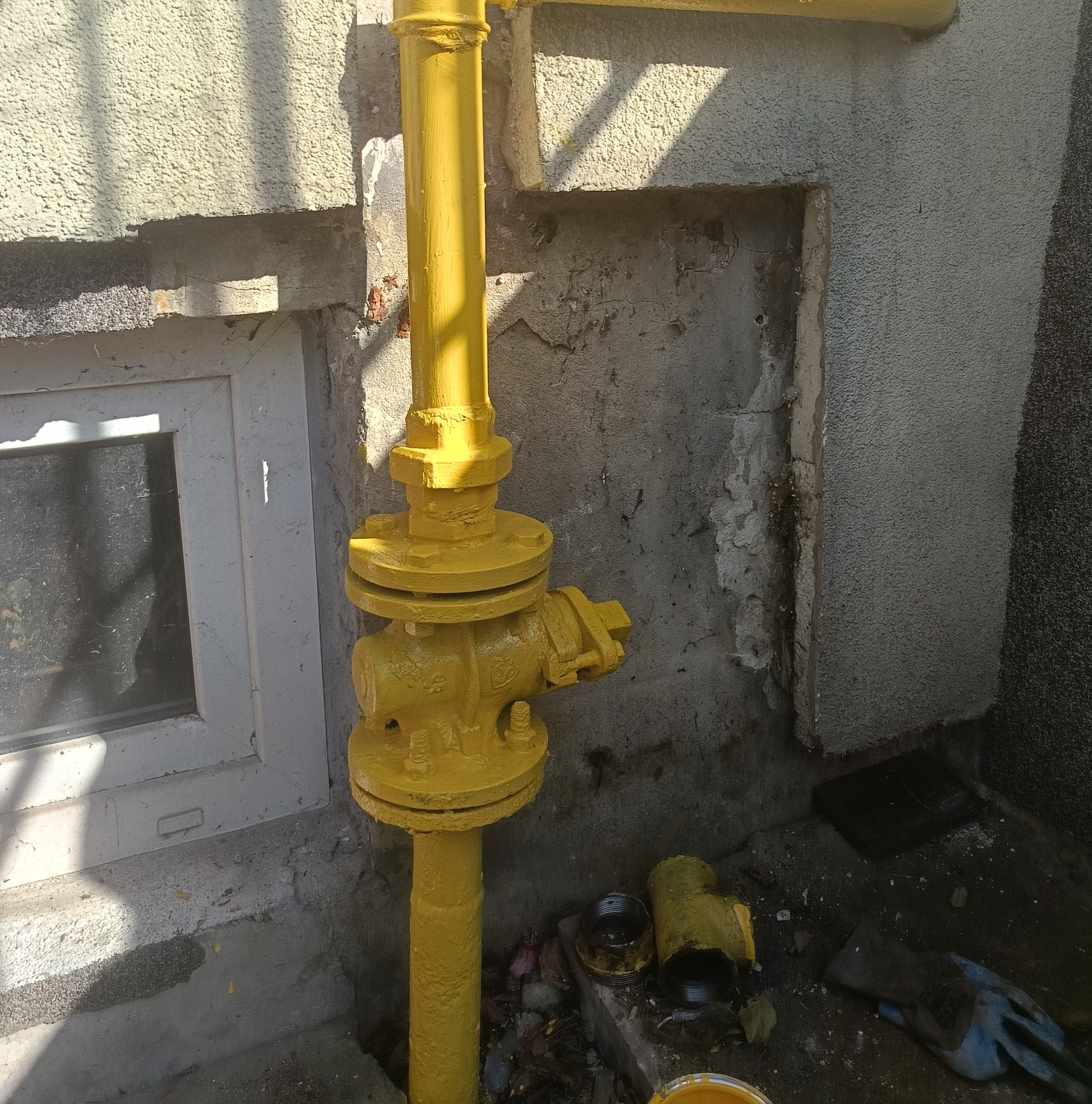 Żółta instalacja gazowa na tle szarej ściany z oknem, świeżo po malowaniu, z widocznymi narzędziami i śladami farby. Instalacja przy budynku.