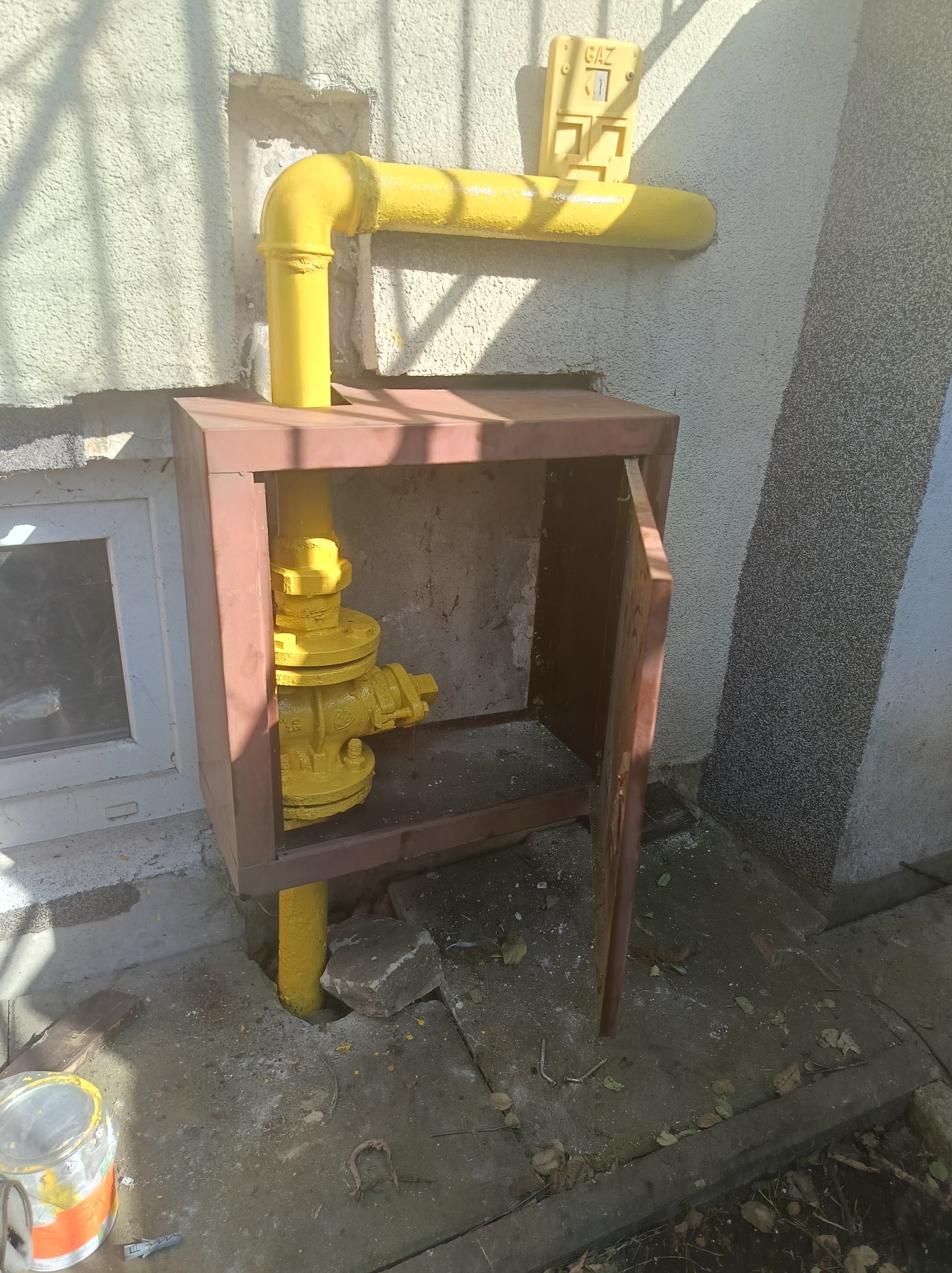 Żółta instalacja gazowa z zaworem w brązowej obudowie przy ścianie budynku. Widoczny licznik gazowy i puszka farby. Otwarta obudowa zaworu gazowego.