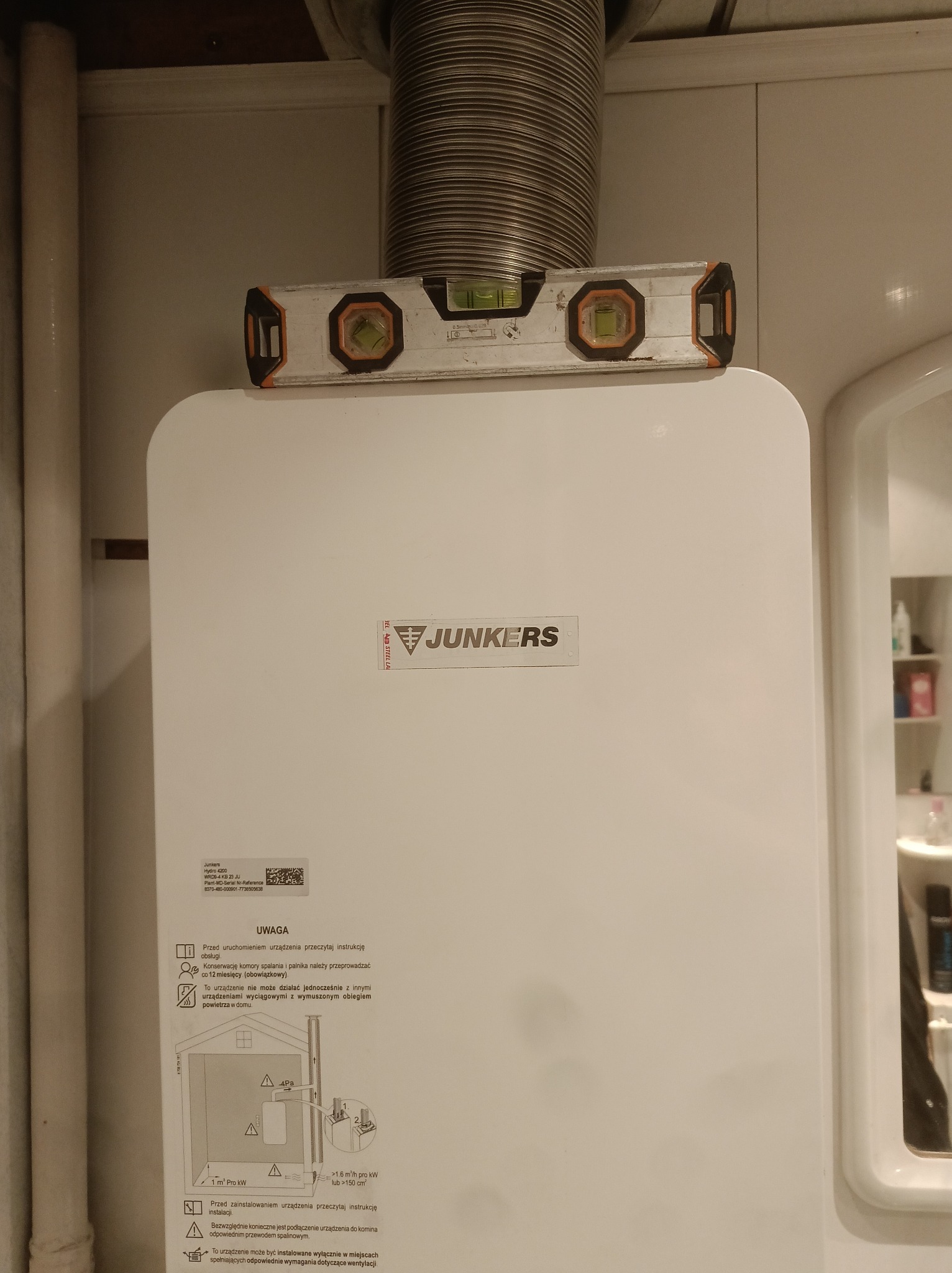 Biały piec gazowy Junkers z poziomnicą na górze, podłączony do rury spalinowej. Widoczna naklejka z instrukcjami i schematem instalacji. Ujęcie z bliska.