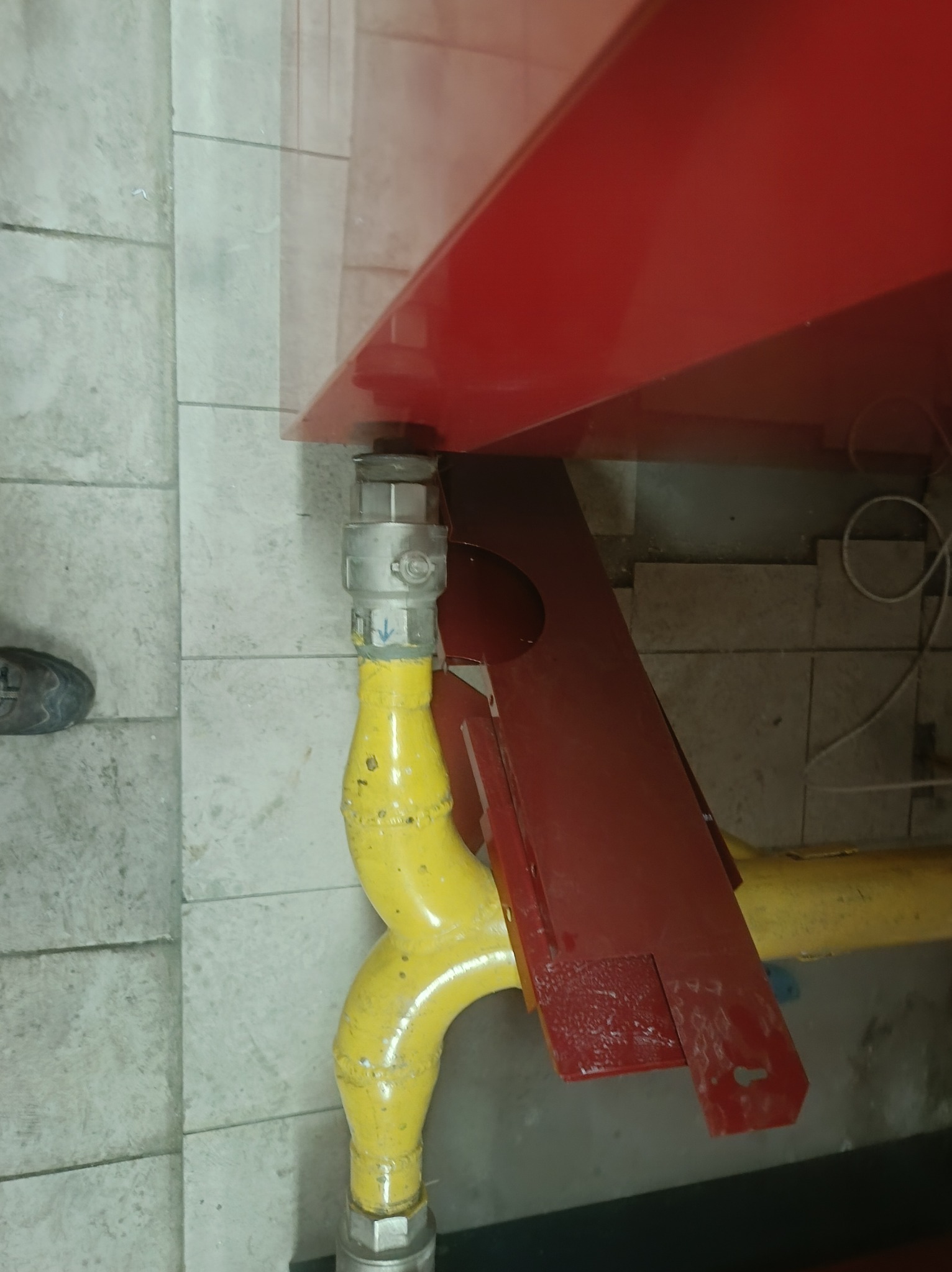 Żółta rura gazowa z zaworem pod czerwonym piecem, widok z dołu, szare płytki w tle, fragment buta w lewym dolnym rogu kadru.