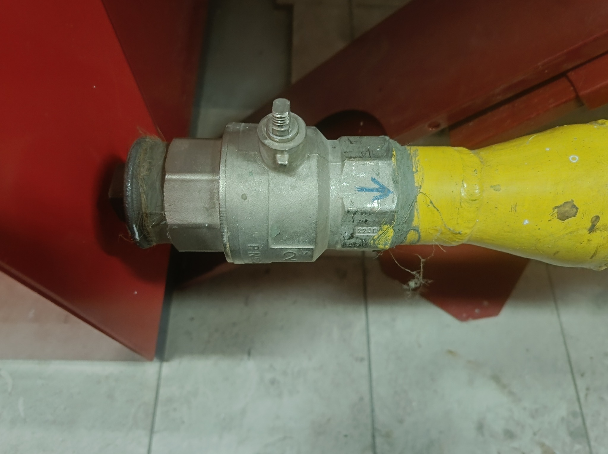 Metalowy zawór z żółtą rurą, widoczny na tle czerwonej obudowy i szarej podłogi. Na zaworze strzałka i oznaczenia. Widoczne ślady użytkowania.