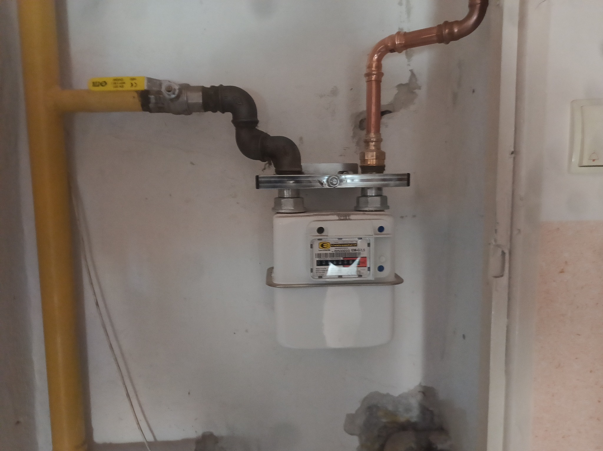 Zamontowany gazomierz z żółtą rurą doprowadzającą gaz i miedzianą rurą odprowadzającą, szara ściana w tle. Widoczne złącza i elementy mocujące.