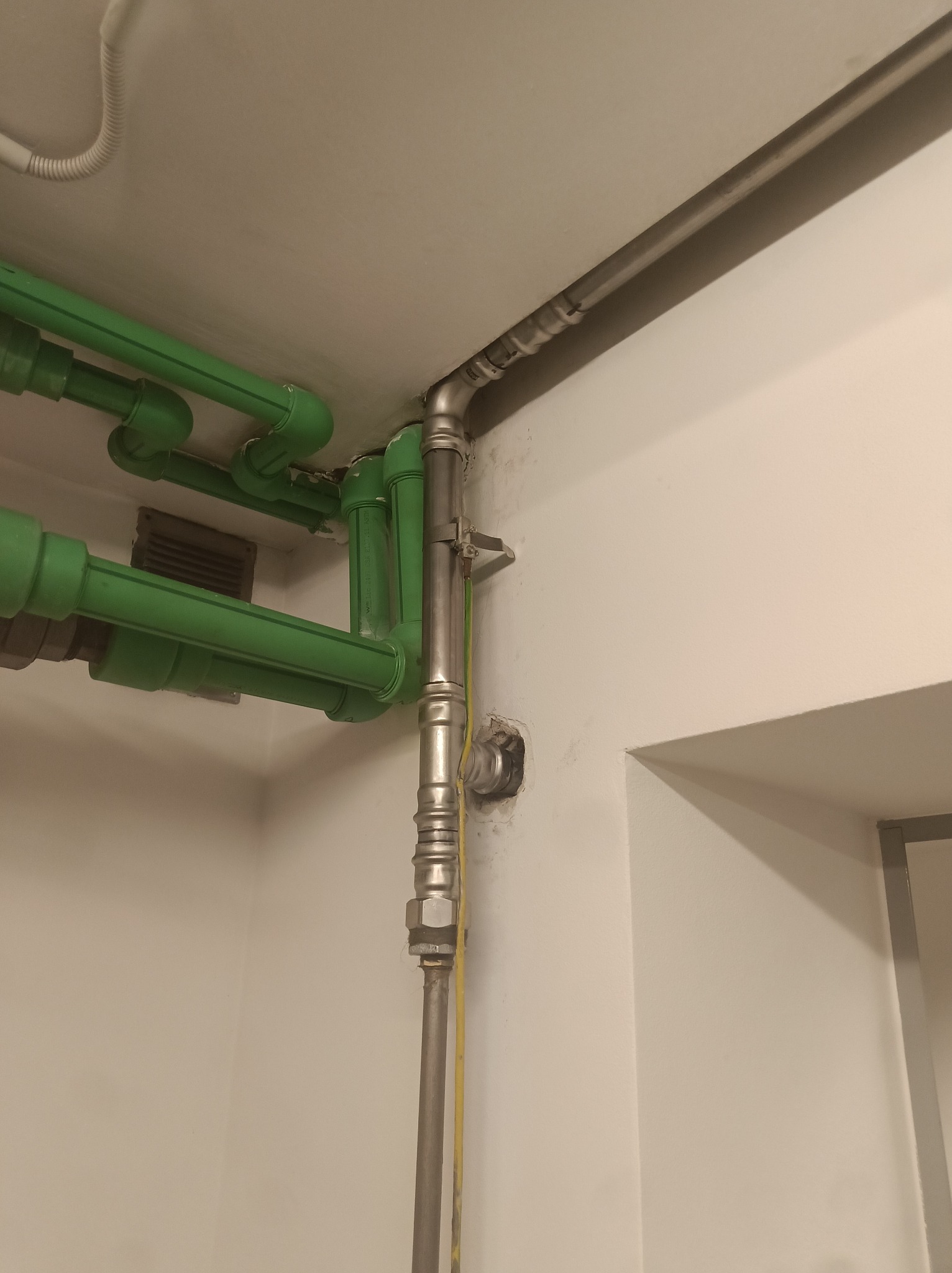 Instalacja wodna i gazowa: zielone rury PP i metalowa rura z żółtym kablem uziemiającym przy ścianie i suficie. Widoczny otwór w ścianie.