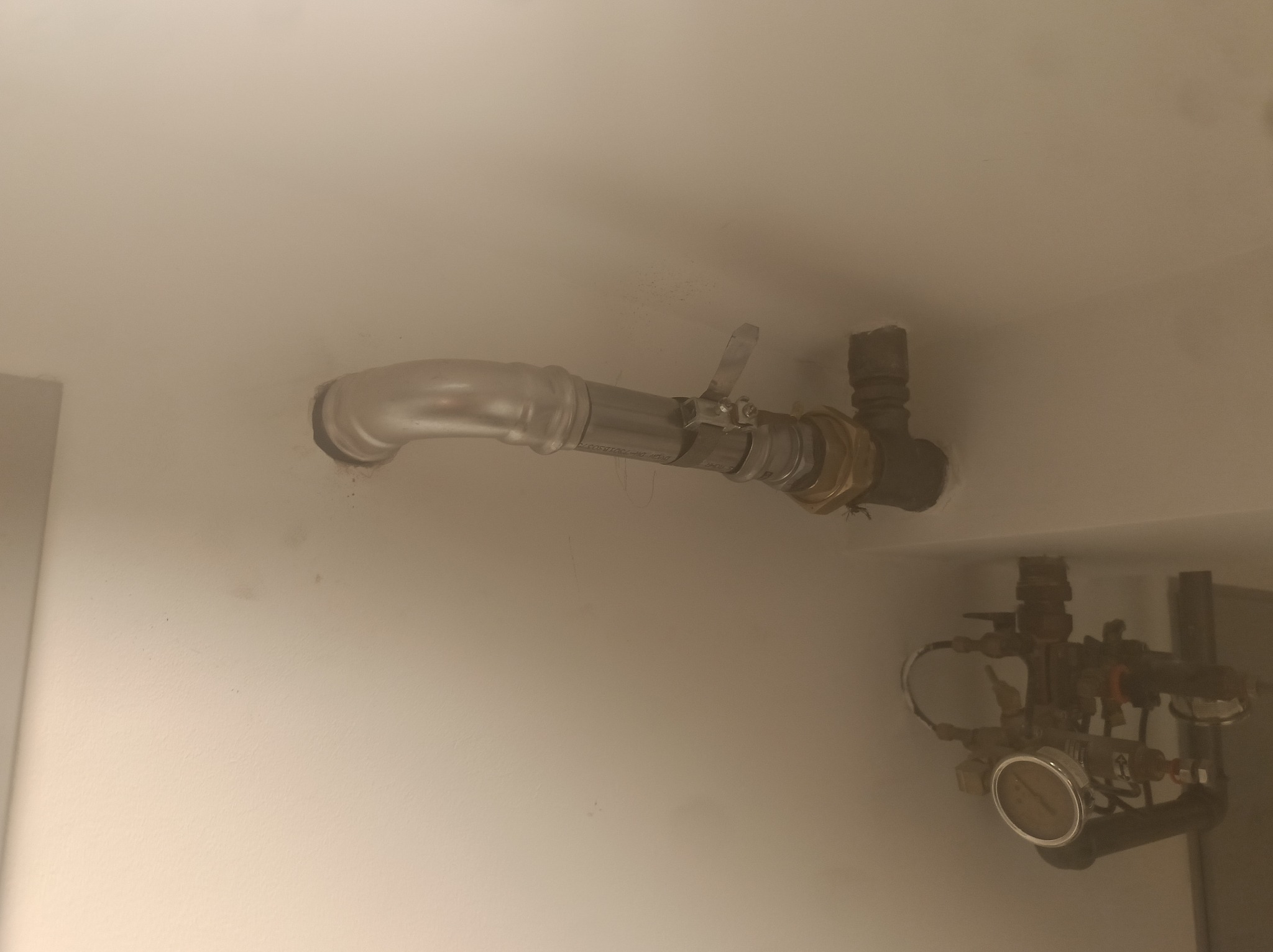 Metalowa rura instalacyjna, z zaworem i złączkami, zamontowana na jasnej ścianie. Widoczny manometr i fragment instalacji w tle.