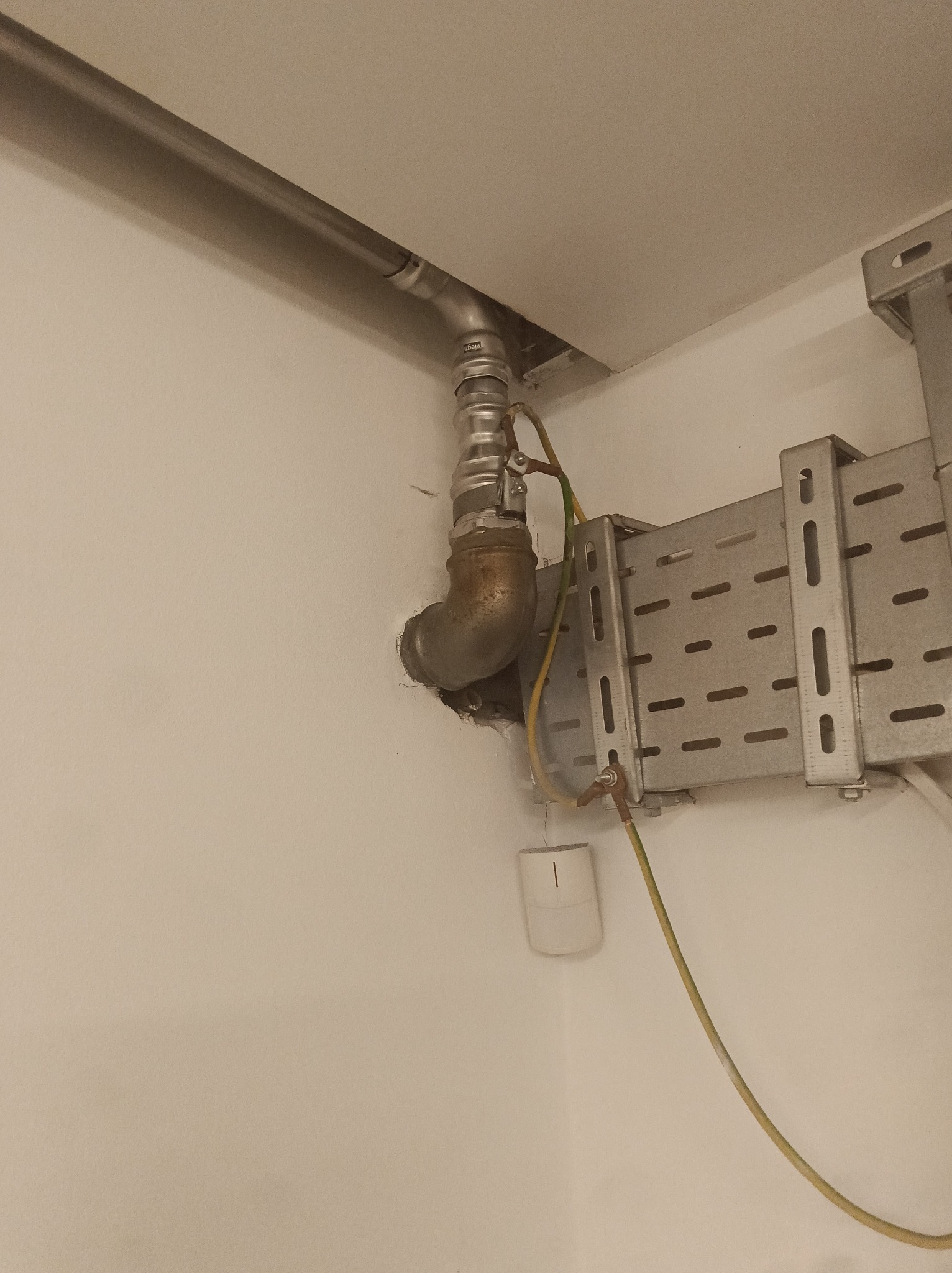 Metalowa rura z kolankiem i kablem uziemiającym wychodząca ze ściany, zamocowana do metalowej konstrukcji wsporczej. Widoczne elementy instalacji.