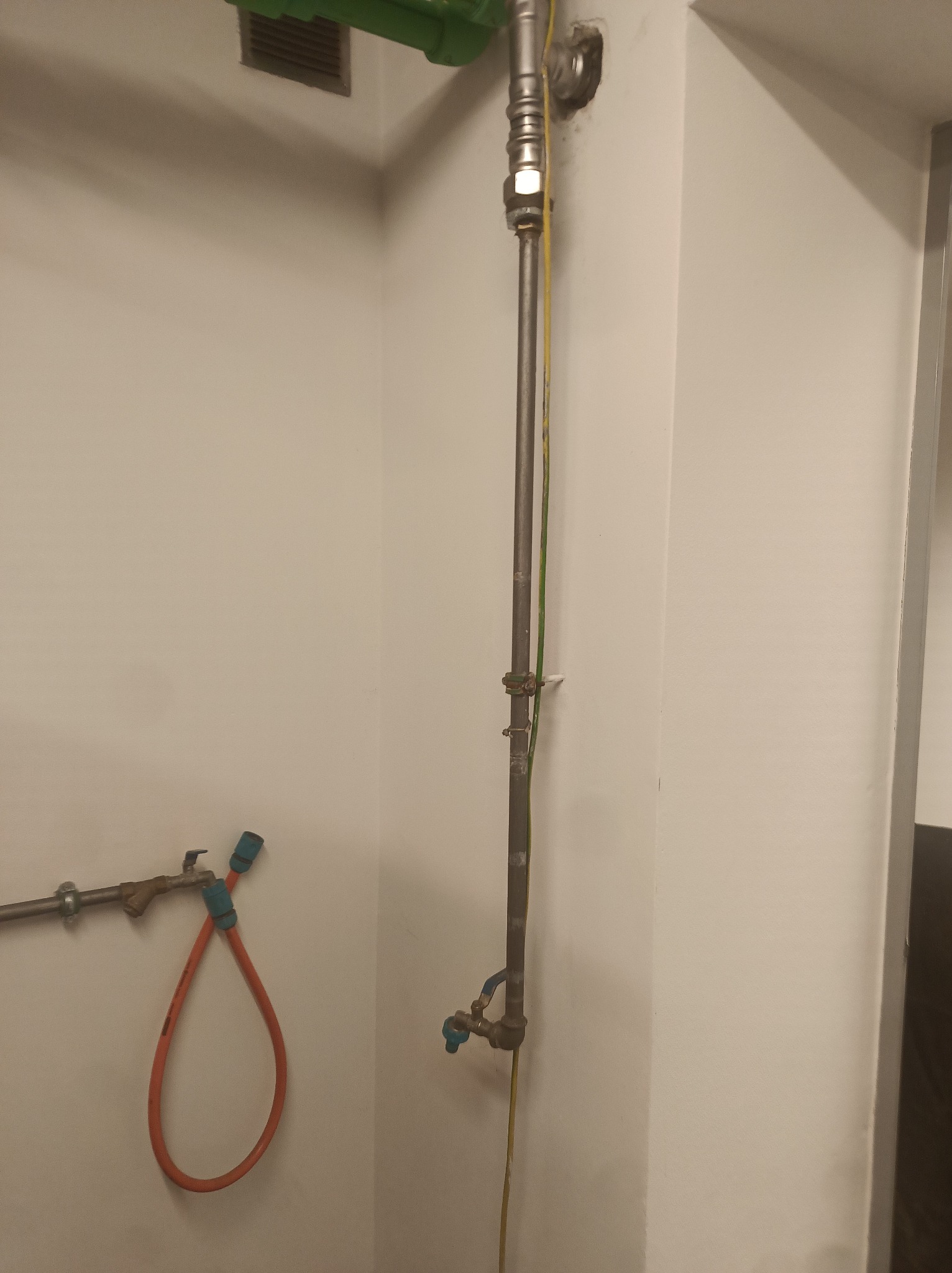 Szara rura gazowa z żółto-zielonym przewodem uziemiającym, biegnąca wzdłuż białej ściany, z kranem na dole i zielonym filtrem u góry. Widoczny wąż gazowy.