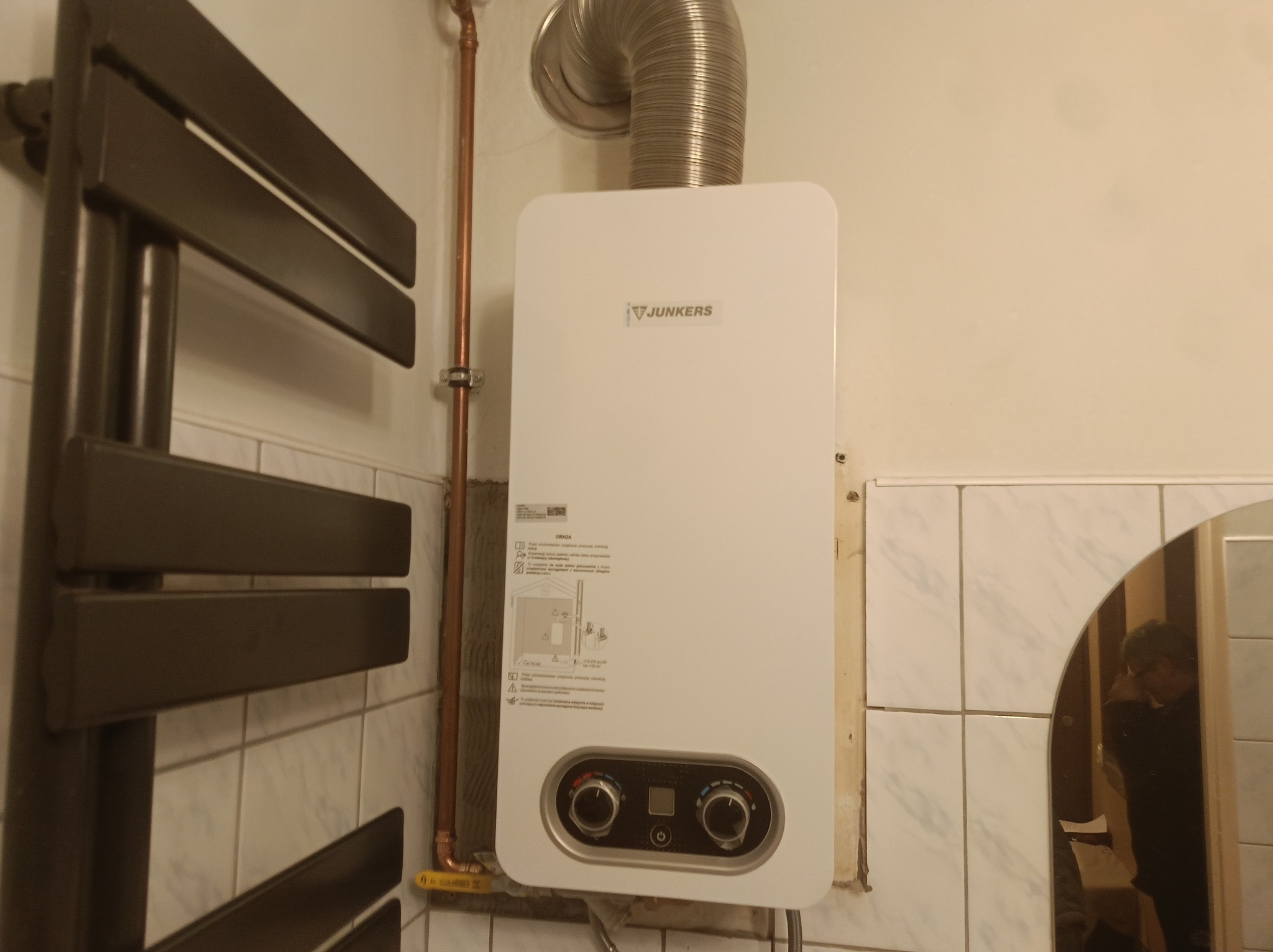 Biały piec gazowy Junkers na ścianie z beżowymi płytkami, obok grzejnik drabinkowy i rura miedziana. Widoczne elementy instalacji i odbicie osoby w lustrze.