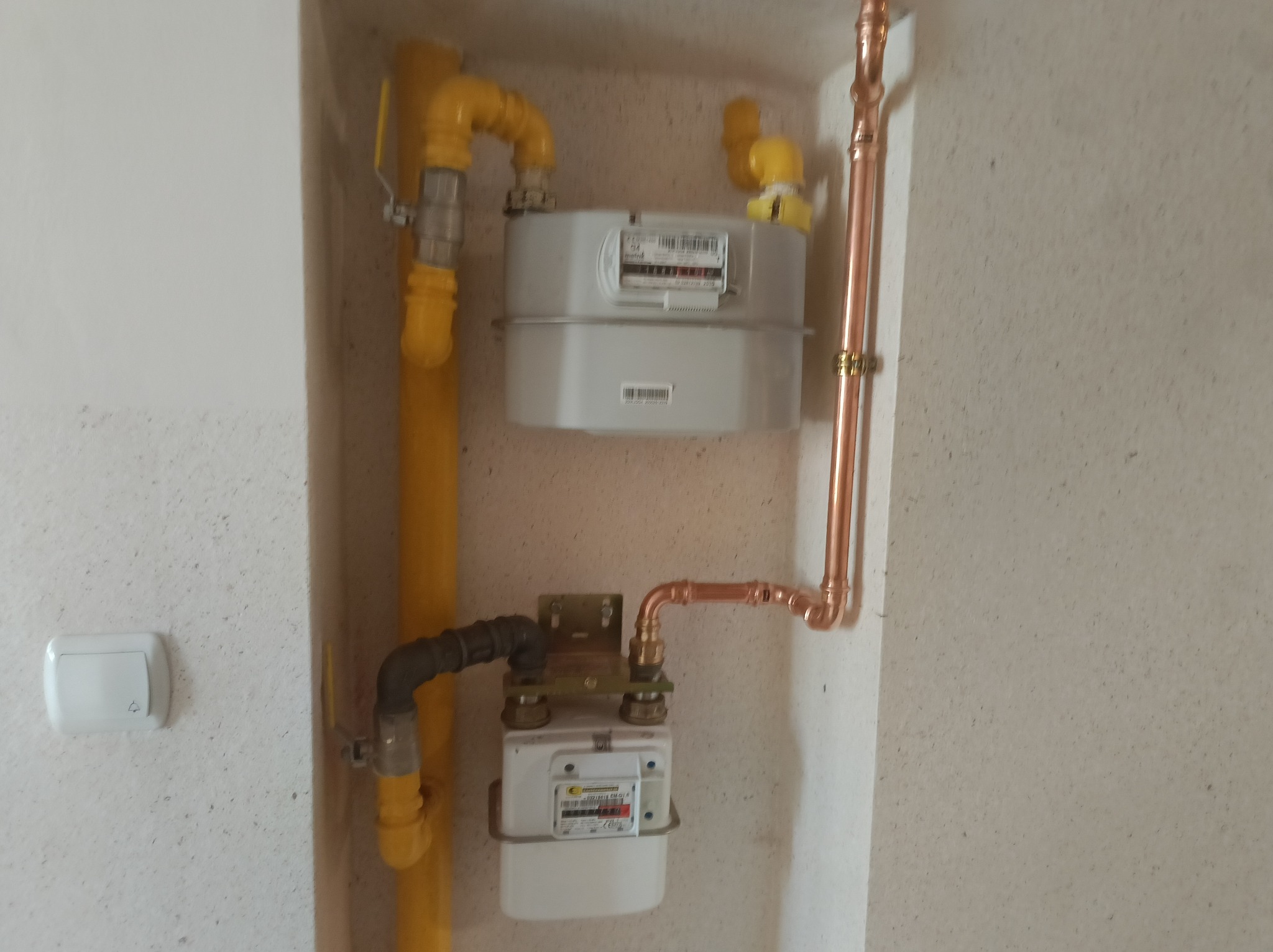 Dwa gazomierze wnęce: górny - nowy, szary, dolny - starszy. Żółte rury gazowe i miedziane rury. Biały dzwonek po lewej. Instalacja gazowa.