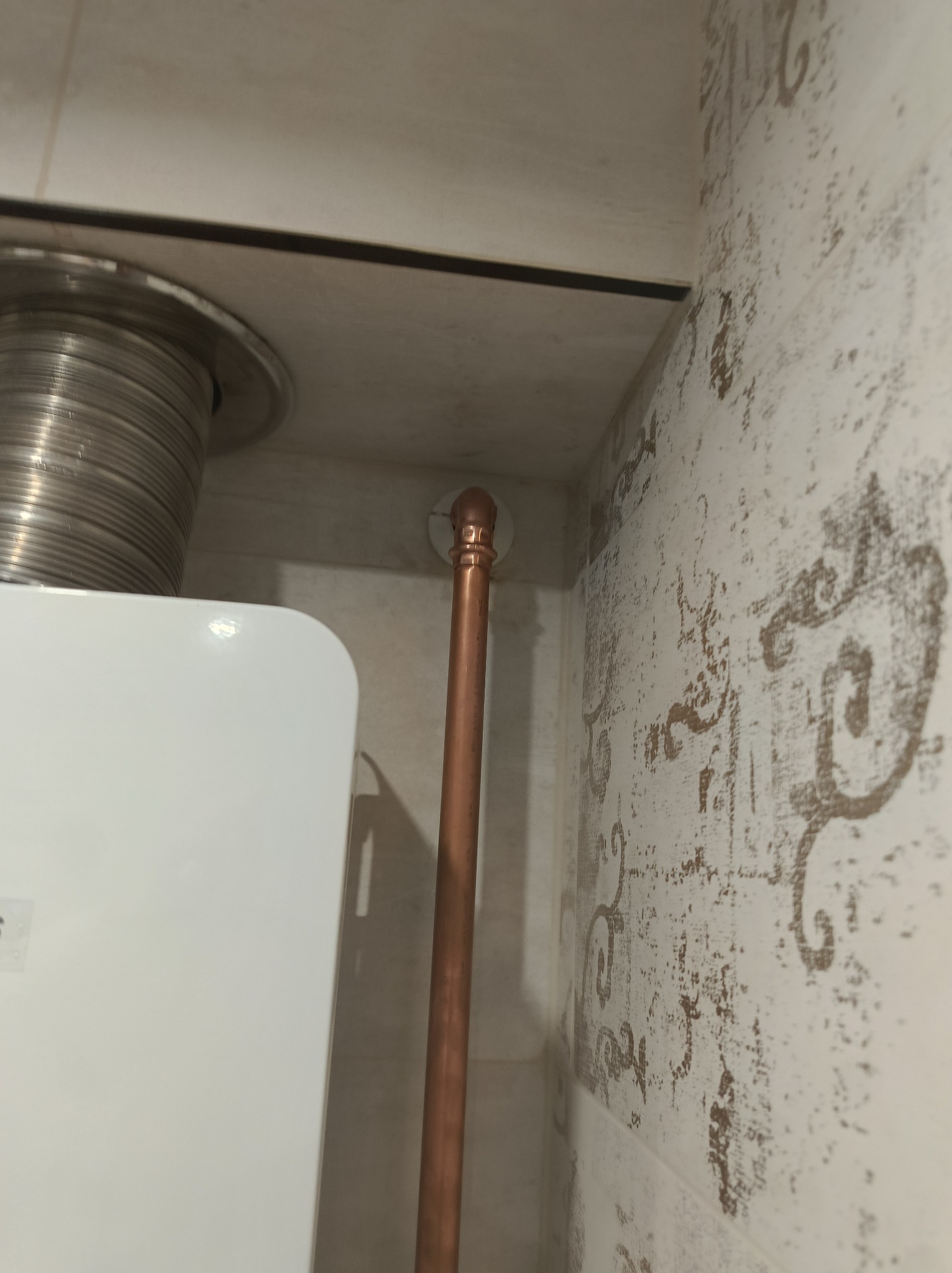 Biały piec gazowy z odprowadzeniem spalin i miedzianą rurą na tle wzorzystych, kremowych płytek. Ujęcie z perspektywy, widoczne elementy instalacji.