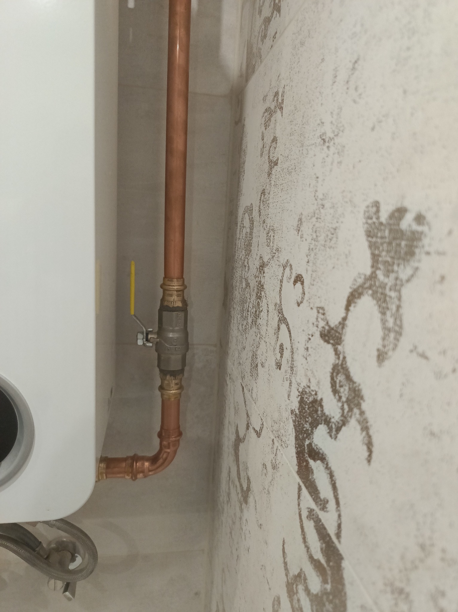 Biały piec gazowy z miedzianą instalacją, zaworem z żółtą dźwignią i elastycznym wężem, na tle ściany z wzorem w łazience. Detal instalacji grzewczej.