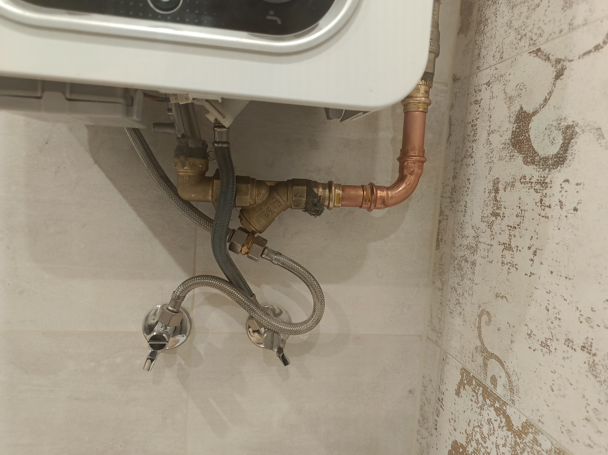 Biały piec gazowy z miedzianymi rurami i metalowymi wężami podłączony do ściany wykończonej płytkami w odcieniach beżu i brązu.