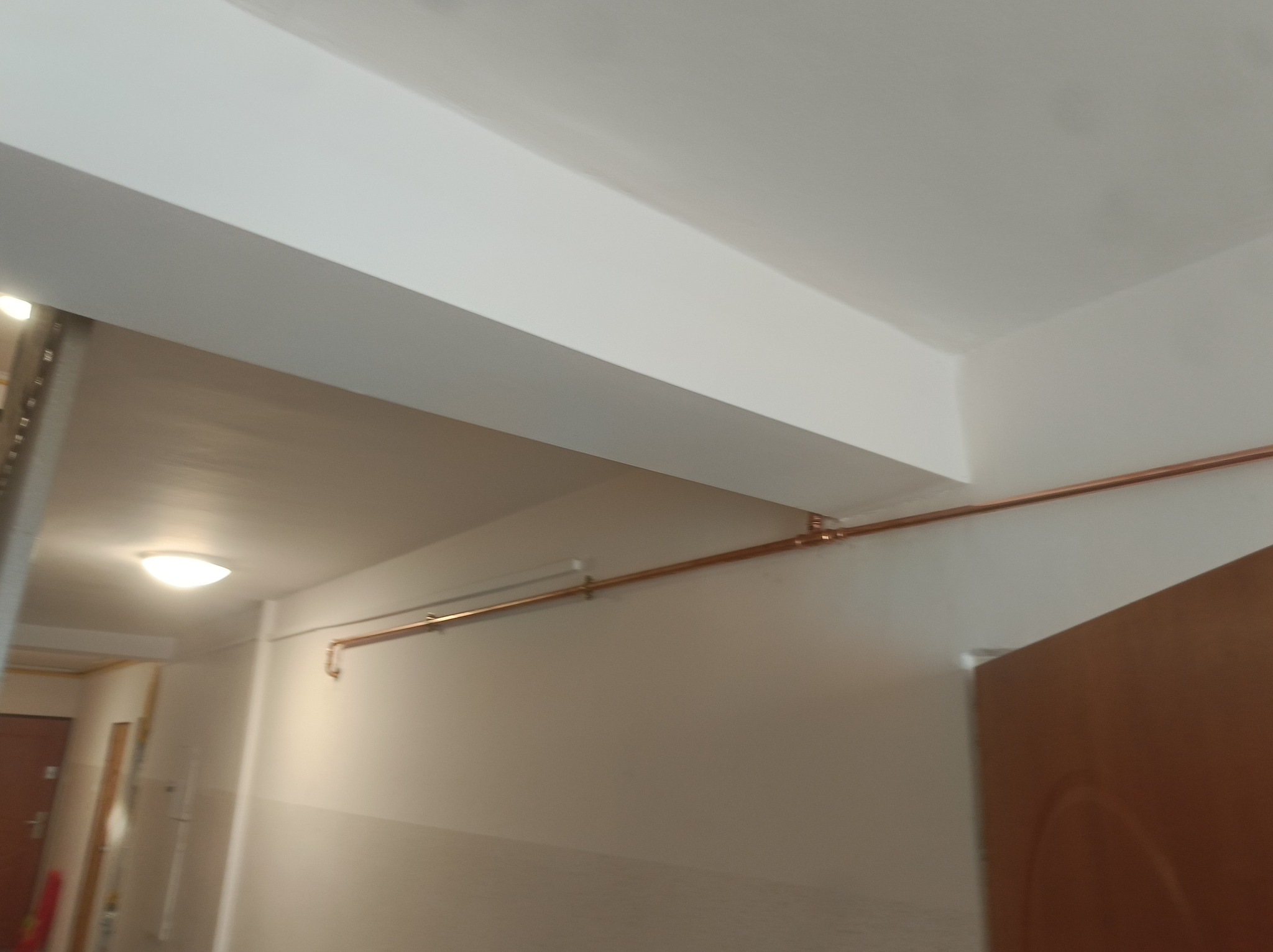 Miedziana rura instalacji biegnąca wzdłuż ściany korytarza, zamocowana do sufitu. Widoczne łączenia rur i elementy mocujące.