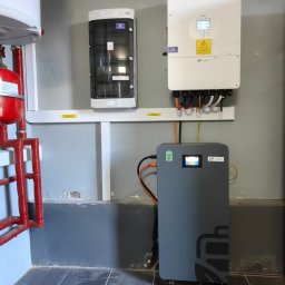 ON SUN - Szara bateria magazynu energii MAKO na kółkach, połączona z białym inwerterem Daye i skrzynką elektryczną na szarej ścianie z instalacją CO.