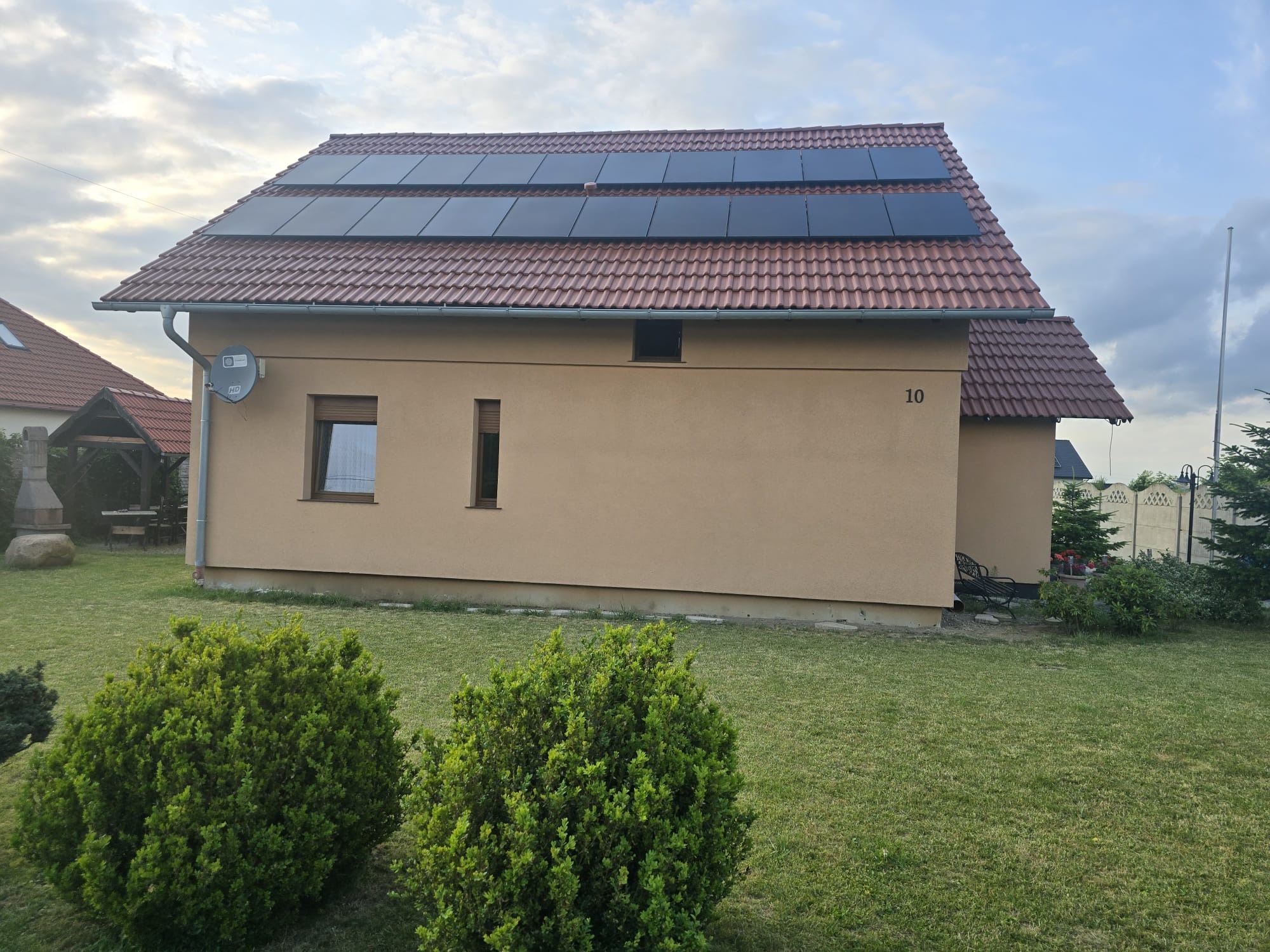 Dom jednorodzinny z panelami słonecznymi na dachu pokrytym czerwoną dachówką. Widoczny numer domu '10', antena satelitarna i bujna zieleń w ogrodzie.