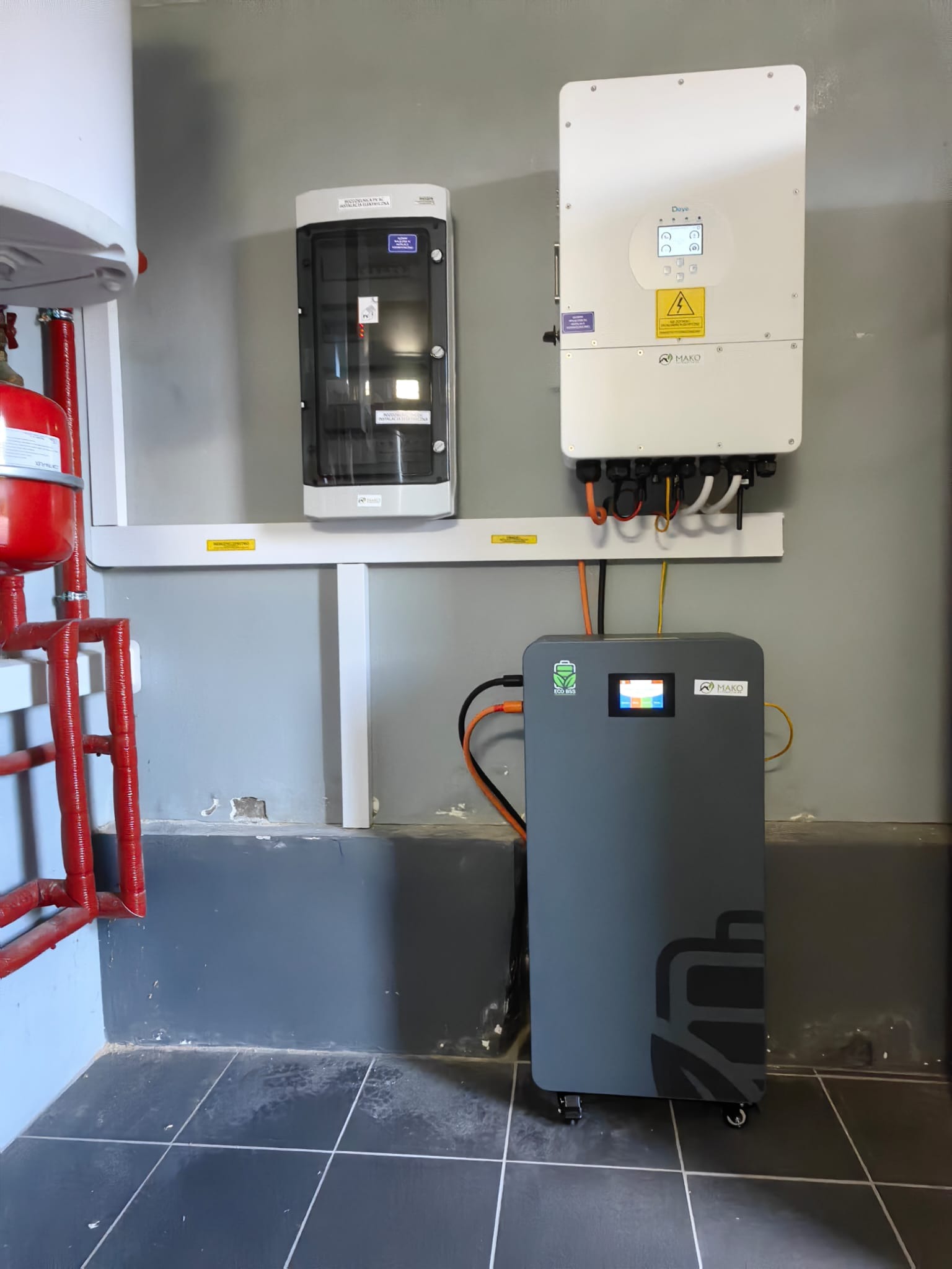 Szara bateria magazynu energii MAKO na kółkach, połączona z białym inwerterem Daye i skrzynką elektryczną na szarej ścianie z instalacją CO.