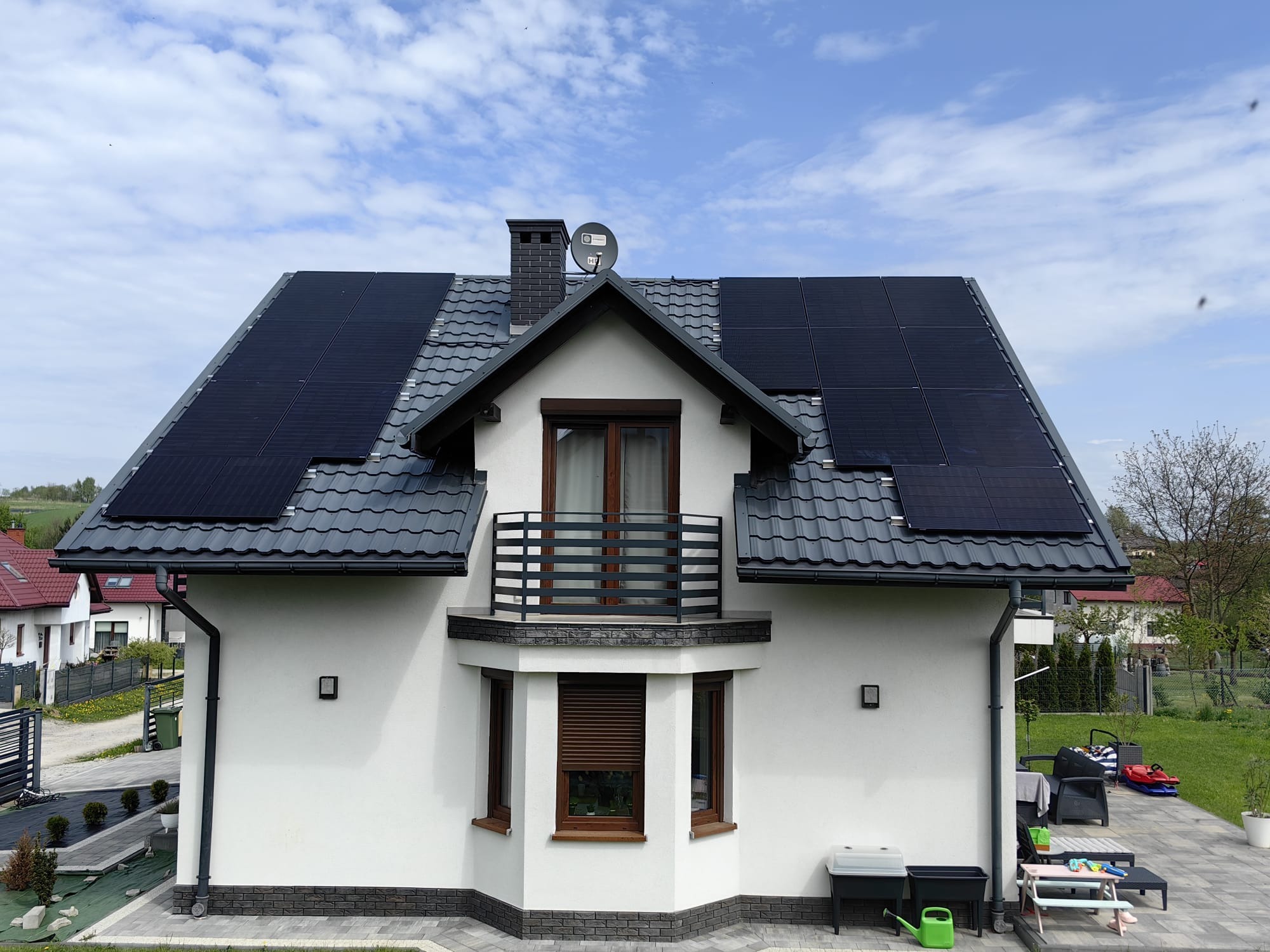 Dom jednorodzinny z szarym dachem i panelami słonecznymi. Ujęcie z poziomu ogrodu, widoczny balkon i elementy małej architektury.