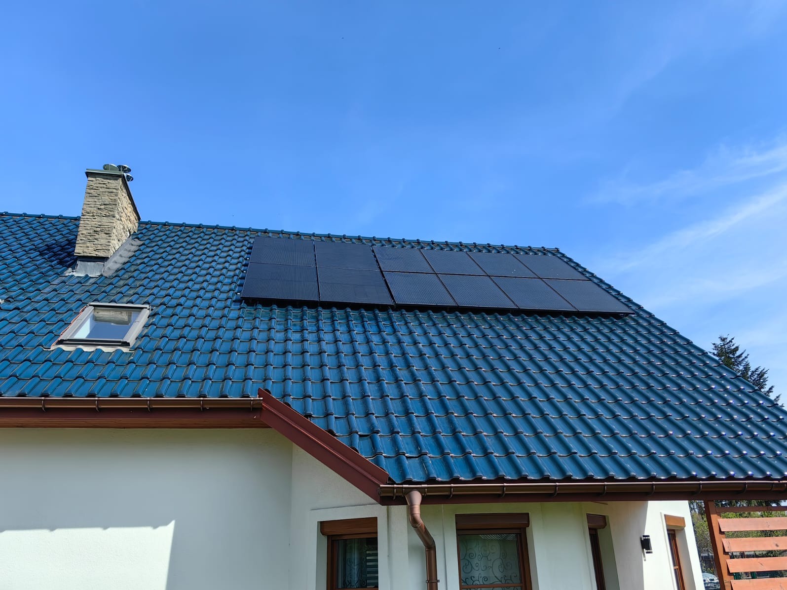 Dom z niebieską dachówką i panelami fotowoltaicznymi. Widok z dołu, jasne niebo. Dachówka błyszcząca, widoczne okno dachowe i komin.