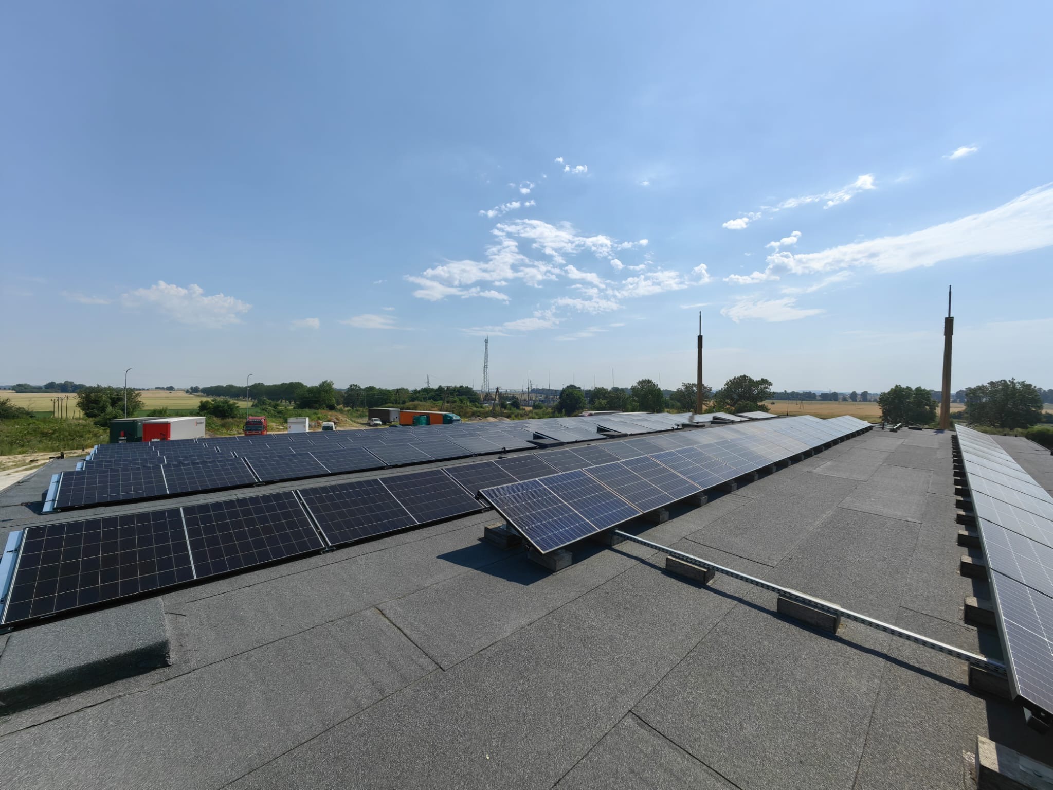 Rząd paneli fotowoltaicznych na płaskim dachu pokrytym papą, w tle błękitne niebo i zielone pola. Widok z góry, w oddali kominy.