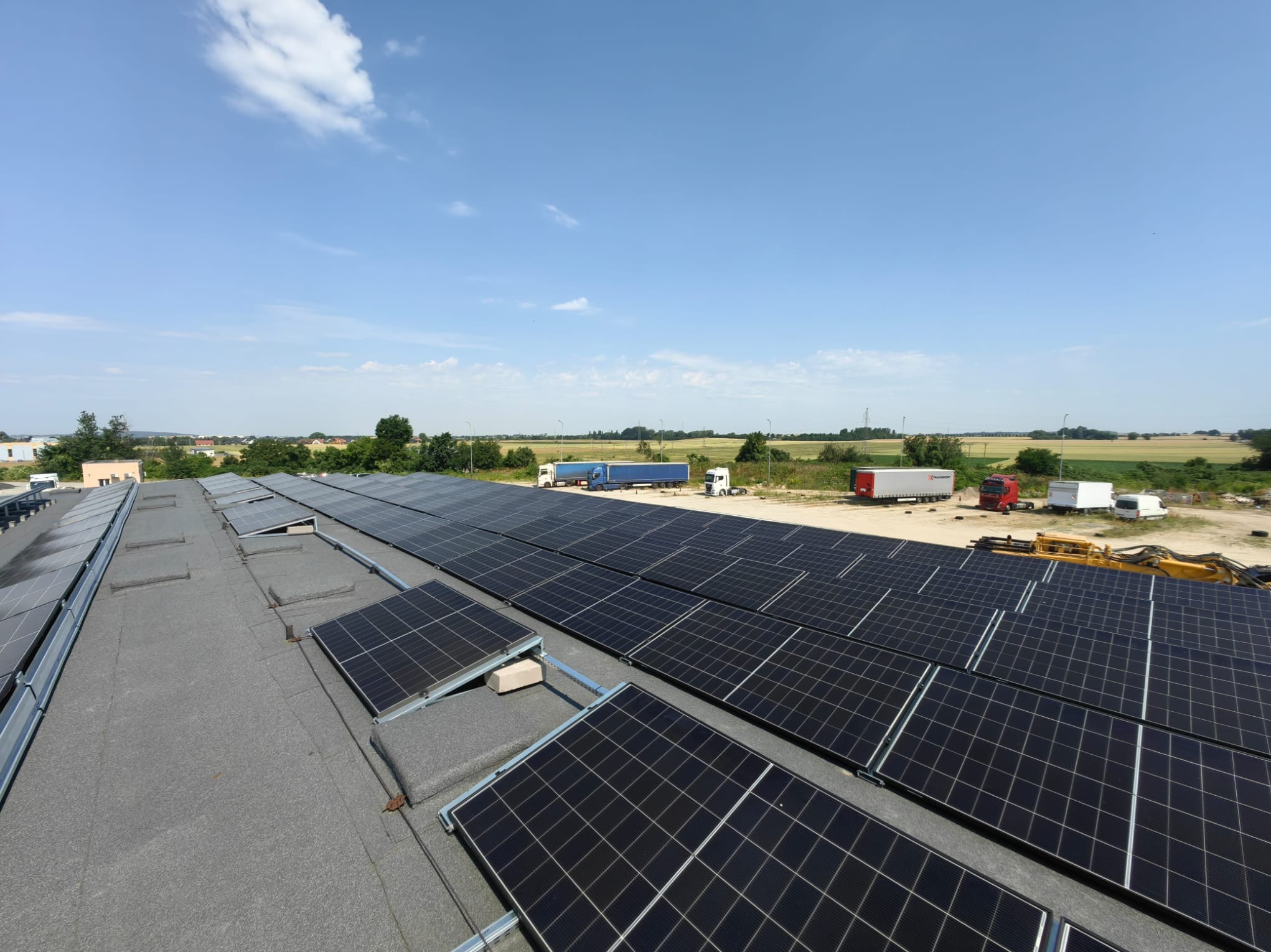 Instalacja fotowoltaiczna na dachu przemysłowym z widokiem na zaparkowane ciężarówki i pola w tle. Panele ułożone równo, widać konstrukcję montażową.